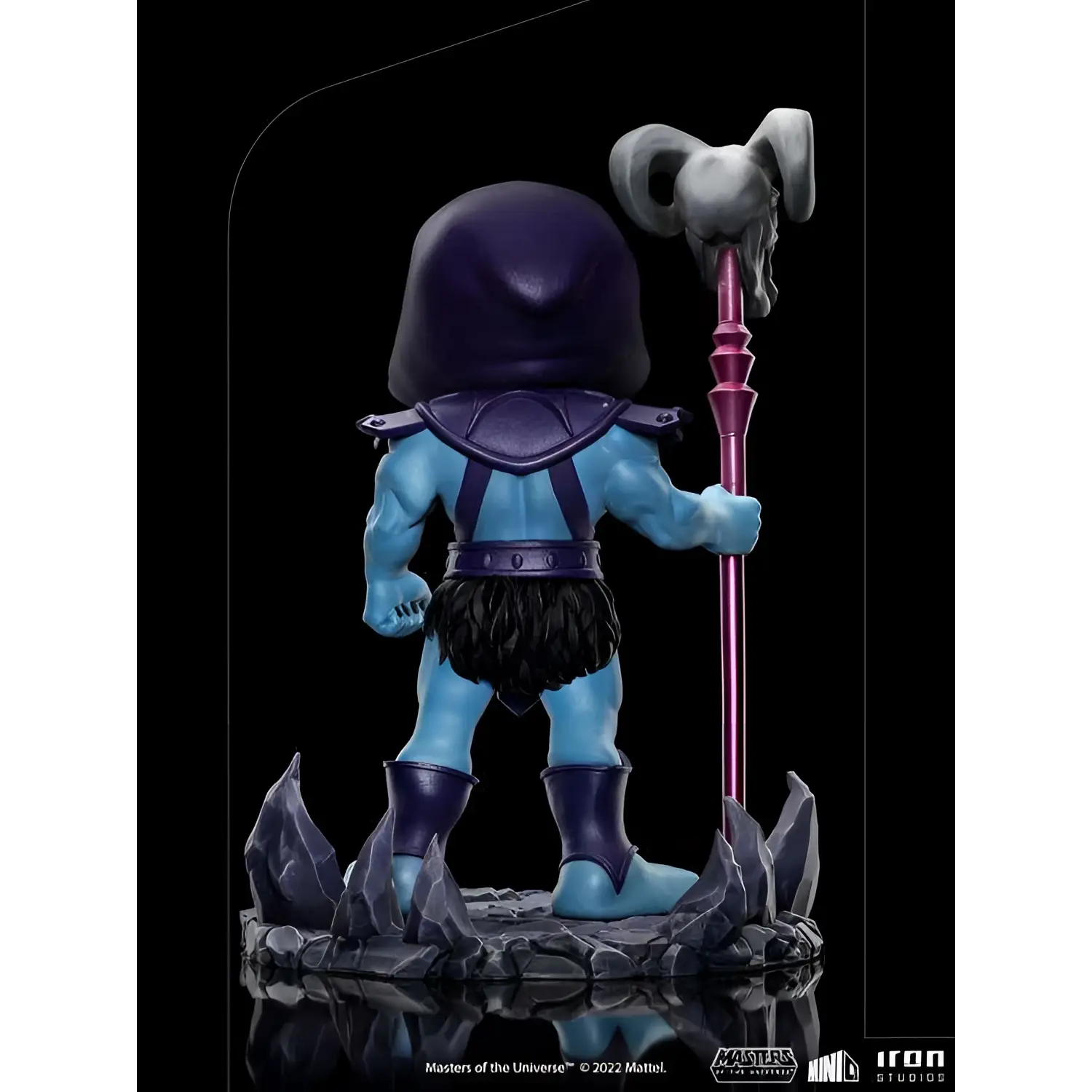 Blå Skeletor-figur i lila huva med rosa stav och grå skalle, på stenbas