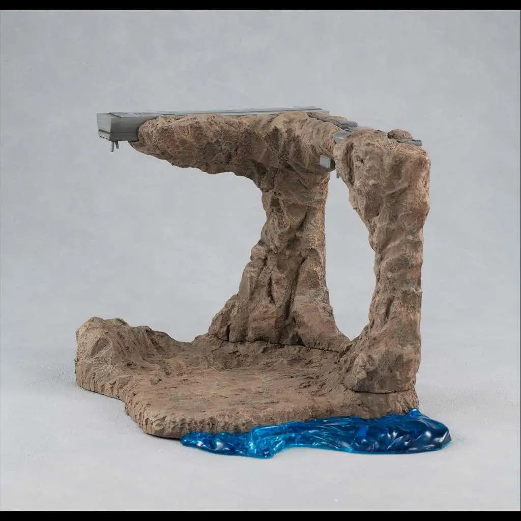 Brun stenig bågebas med blå genomskinlig vätskapool och grå metallkomponenter i Ms Gundam G Structure Tragedy Diorama